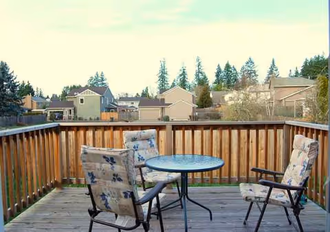 Outdoor wooden deck with a round glass table and cushioned chairs overlooking neighborhood homes.
