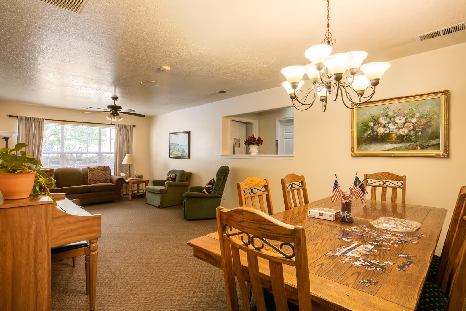 A cozy living and dining area in a senior living facility. The dining area features a wooden table with six chairs, a chandelier overhead, and a partially completed puzzle on the table with small American flags in a vase. The living area has a brown sofa, two green armchairs, a side table with a lamp, a ceiling fan, and a large window with curtains letting in natural light. There is a piano with a potted plant on it and framed paintings on the walls.
