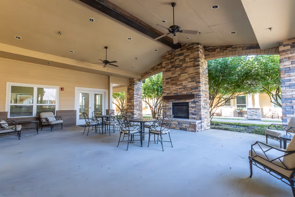 Covered outdoor patio area with stone pillars and a built-in fireplace. The space includes several metal tables with chairs and cushioned seating along the walls. Ceiling fans are mounted on the beige ceiling with recessed lighting. Trees and greenery are visible outside the patio area.