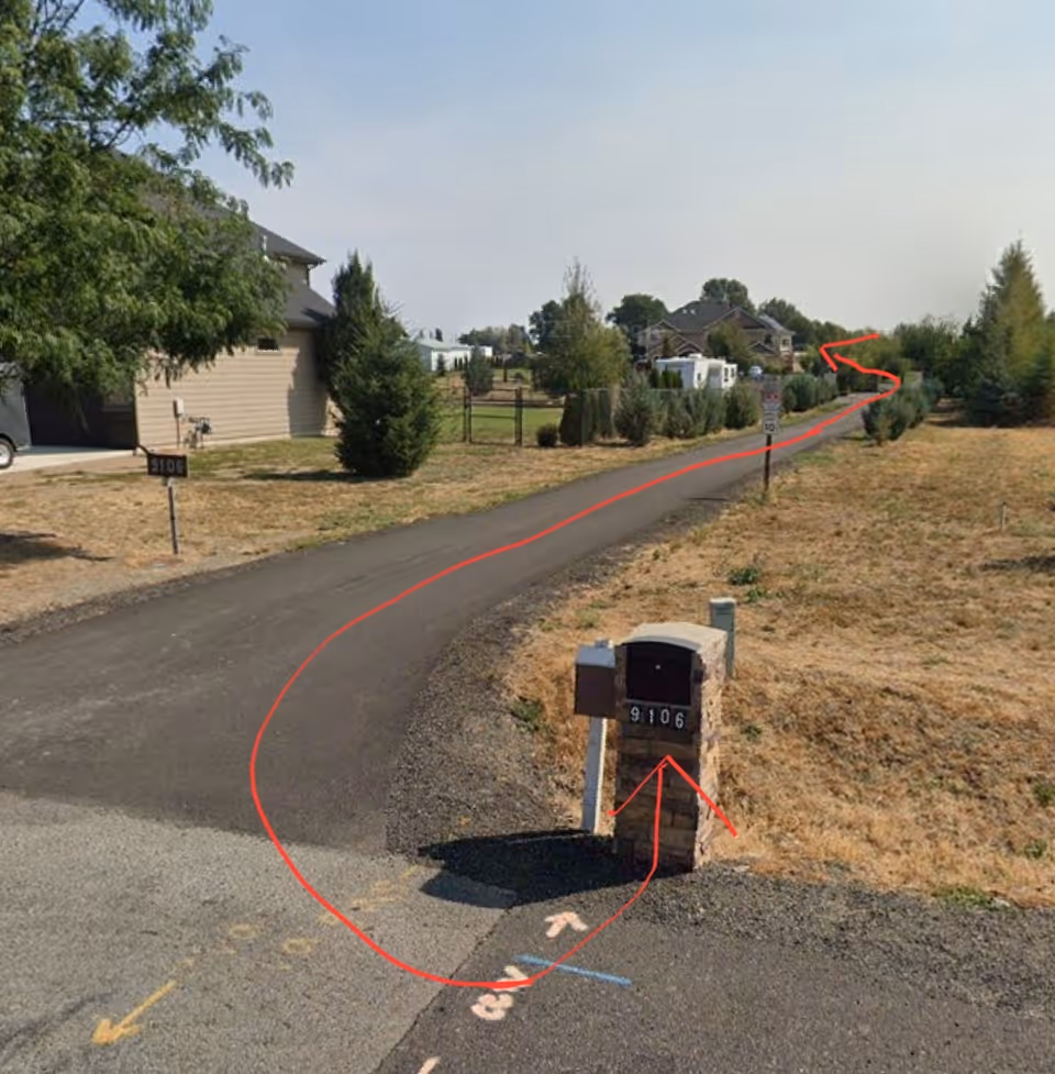 A paved driveway curving through a residential area with dry grass and small trees on either side. There is a brick mailbox with the number 9106 on it near the entrance of the driveway. Houses and more trees are visible in the background under a clear sky.