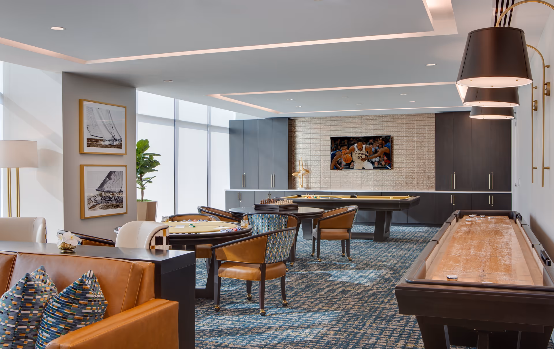 A modern game room with a shuffleboard table, a pool table, and a card table with chairs. The room has a patterned carpet, a leather sofa with colorful pillows, framed pictures of sailboats on the wall, and a flat-screen TV mounted on a textured wall. Large windows with frosted glass allow natural light to fill the space.