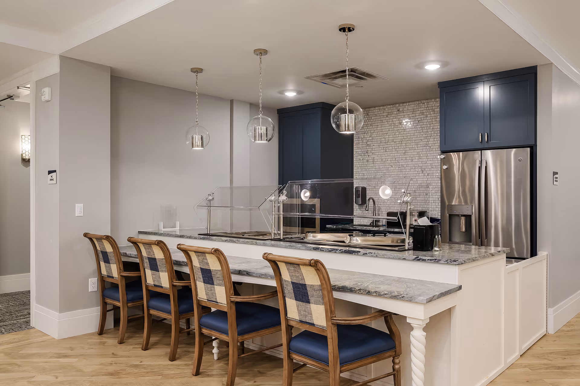Modern kitchen area with a marble countertop island featuring a clear sneeze guard, four wooden chairs with blue and beige plaid cushions, stainless steel refrigerator, blue cabinets, and pendant lights hanging from the ceiling.