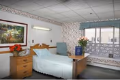 A single hospital-style bed with light blue bedding in a room with floral wallpaper and a large window with a blue valance. Next to the bed is a wooden nightstand with a vase of red flowers and a wall-mounted light above the bed. A wooden overbed table holds a vase of pink flowers and a blue book. There is a light blue armchair near the window and a framed nature painting on the wall.