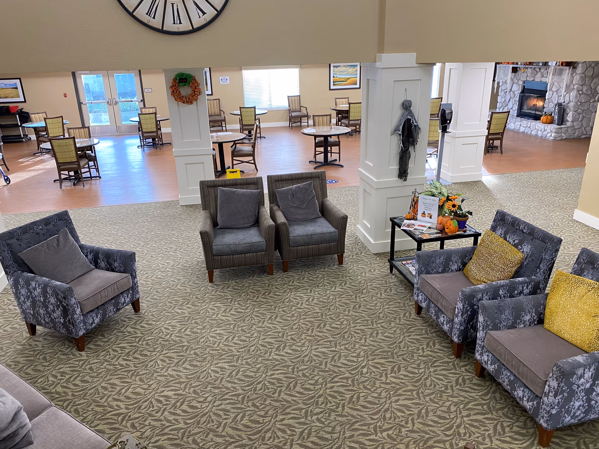 A spacious common area in an assisted living facility featuring several upholstered armchairs with cushions arranged around a small table decorated with fall-themed items. In the background, there are multiple round tables with chairs, large windows letting in natural light, and a stone fireplace with a lit fire. The floor is carpeted in the seating area and transitions to wood flooring in the dining area.