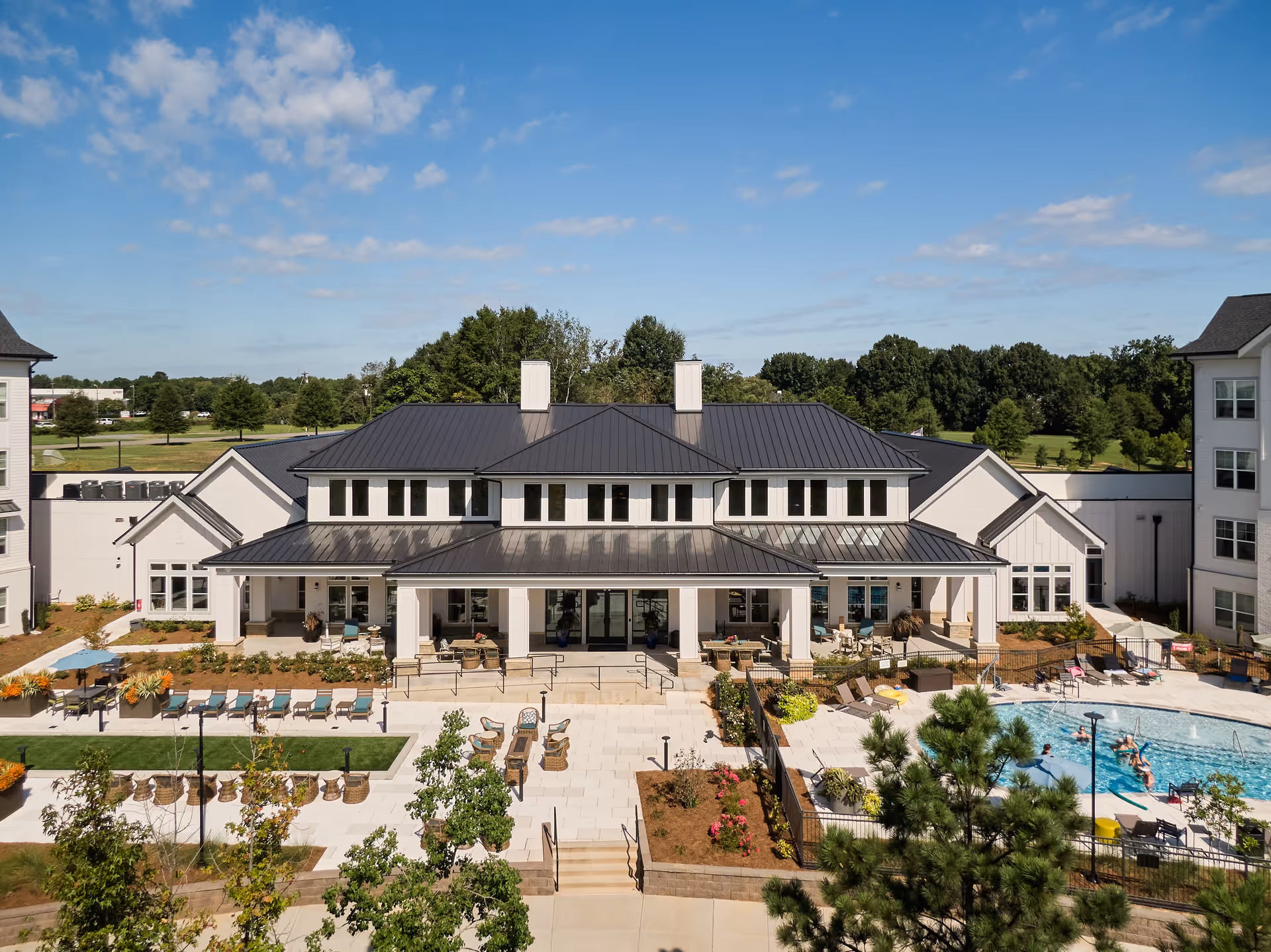 Exterior view of Inspire Royal Park facility showing a large building with a dark metal roof, surrounded by landscaped gardens, outdoor seating areas, and a swimming pool with people enjoying the water under a partly cloudy blue sky.