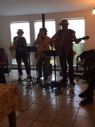 Three musicians performing indoors in front of large windows. One person is playing a guitar, another is playing a wind instrument, and the third is singing into a microphone. The room has tiled floors and some chairs and tables are partially visible.