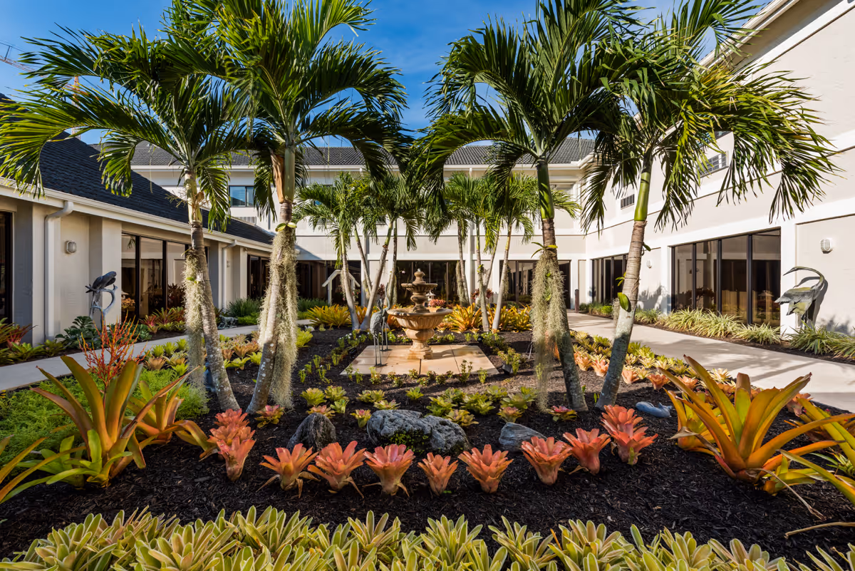 A landscaped courtyard with palm trees, various tropical plants, and a central water fountain surrounded by a two-story building with large windows under a clear blue sky.