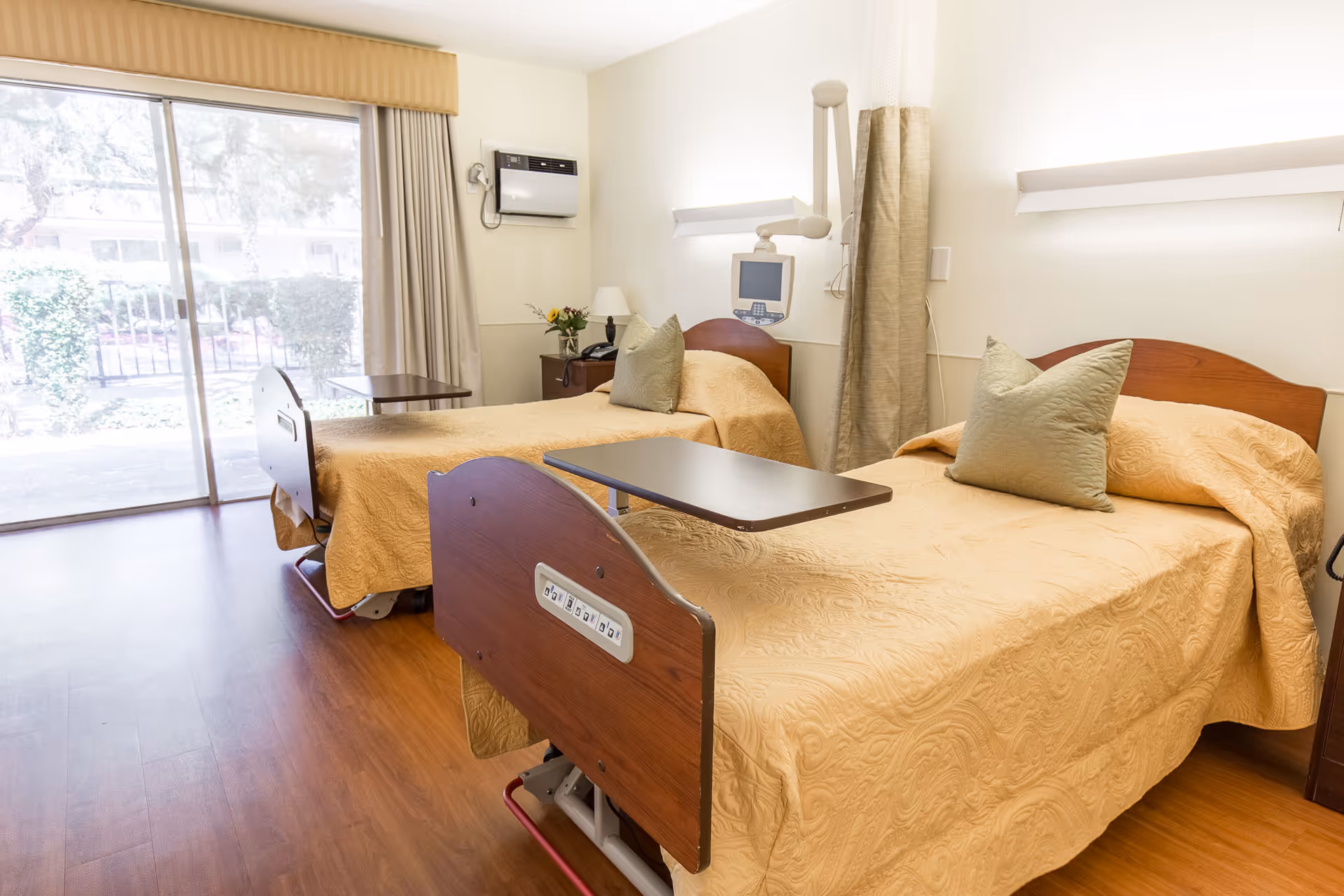 A bright room with two hospital-style beds covered in beige bedspreads, each with a green pillow. There is a small table attached to one bed and a bedside table with a lamp and flowers between the beds. A large sliding glass door with curtains lets in natural light and shows greenery outside. The room has wooden flooring and medical equipment mounted on the wall.
