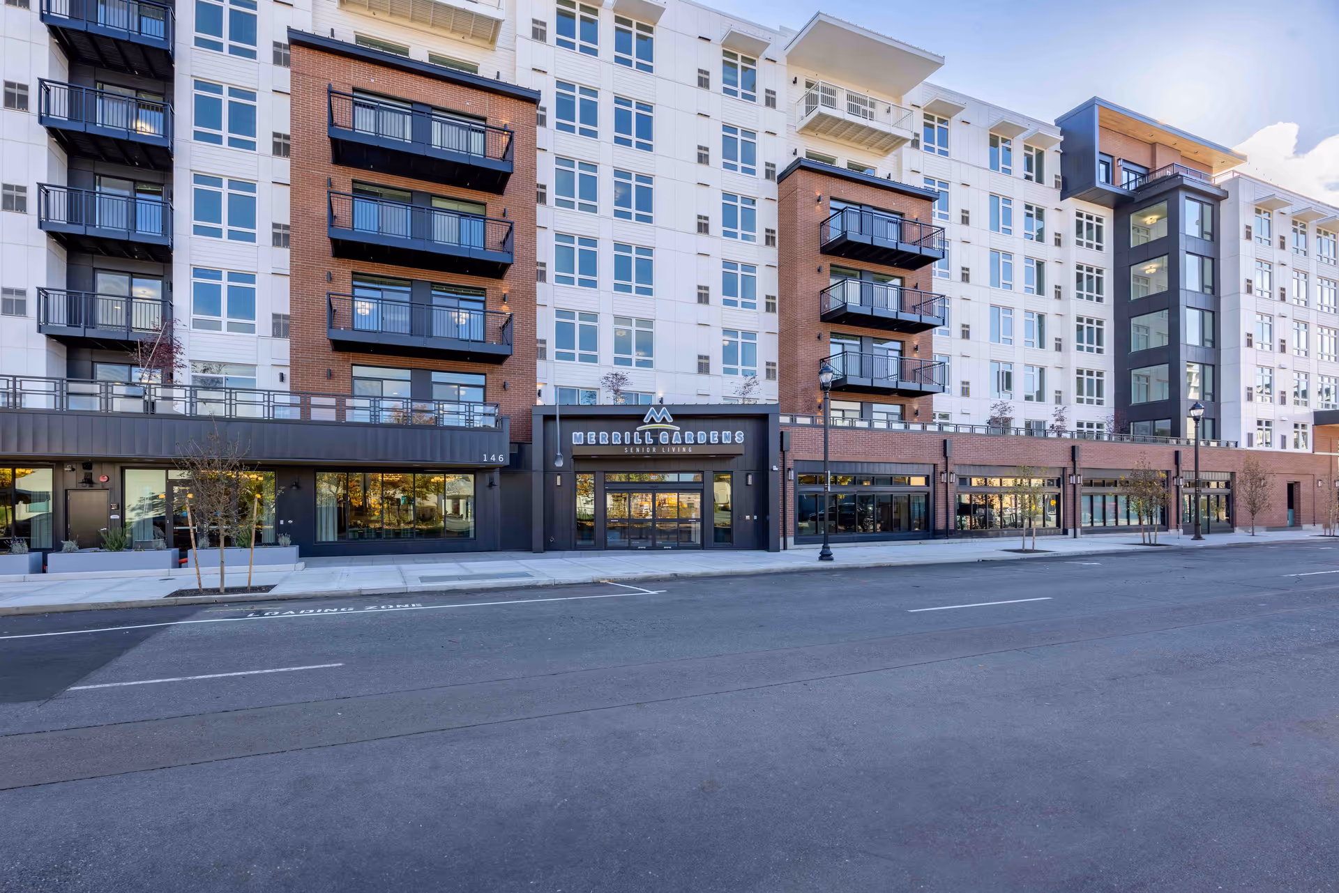 Exterior view of a multi-story senior living facility named Merrill Gardens at Hillsboro, featuring balconies, large windows, and a main entrance with the facility's name above it. The building has a modern design with brick and white paneling, and there is a street with a loading zone in front.