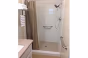 A clean and accessible shower area with a beige shower curtain, grab bars on the wall, a handheld showerhead, and a tiled floor. A countertop with a sink and folded towels on a rack are visible to the left.