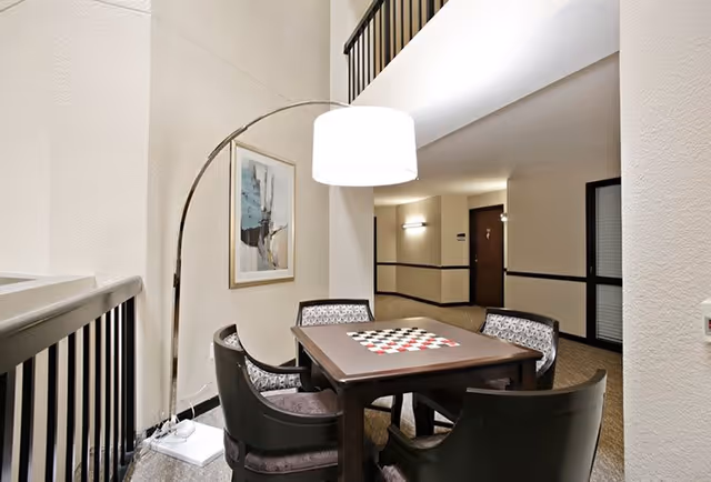 A cozy seating area in a hallway with a square table featuring a checkerboard pattern on top, surrounded by four cushioned chairs. A tall arc floor lamp with a white lampshade illuminates the space. The walls are light-colored with a framed abstract painting, and there is a railing on the left side overlooking a lower level.