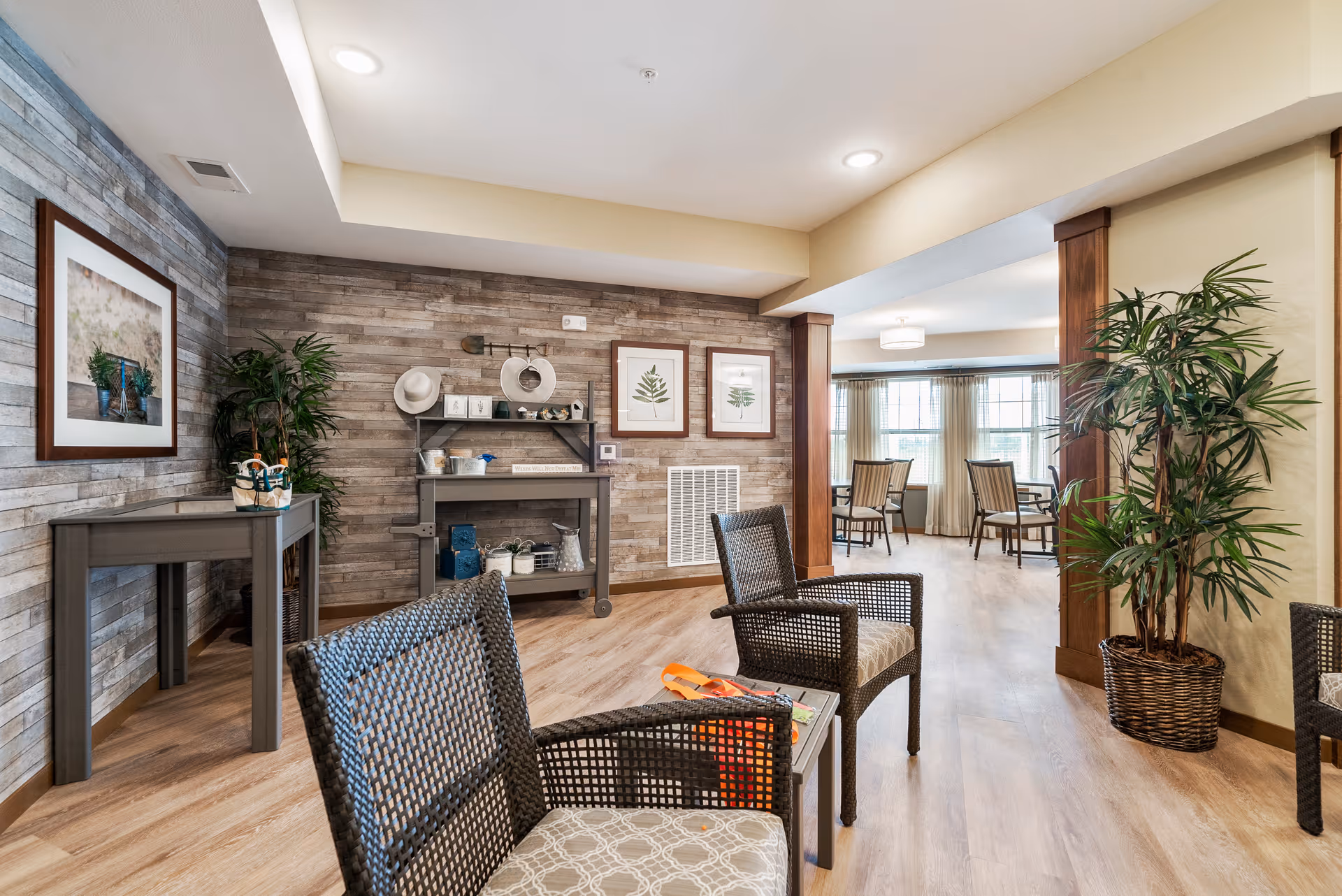 A cozy senior living common area with wicker chairs and a small table in the foreground. The walls have a wood panel design with framed botanical prints and decorative items on shelves. Large windows with sheer curtains allow natural light into the adjacent dining area with tables and chairs. Potted plants add greenery to the space.