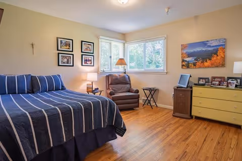 Well-lit bedroom with hardwood floors, a bed with blue striped bedding, a recliner by corner windows, and a dresser with framed photos.