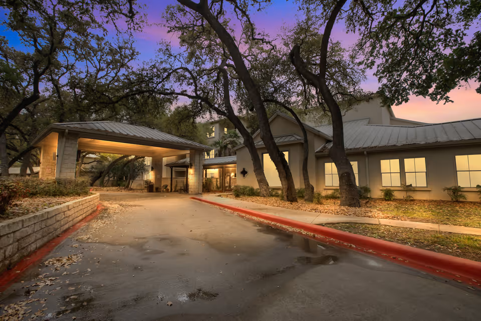 Exterior view of The Inn at Los Patios facility at dusk, showing a driveway leading to a covered entrance with warm lighting. The building is surrounded by trees and landscaping, with a purple and orange sky in the background.