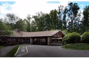 Exterior view of a single-story nursing and rehabilitation center building surrounded by trees and shrubs, with a driveway leading to the entrance under a covered drop-off area.