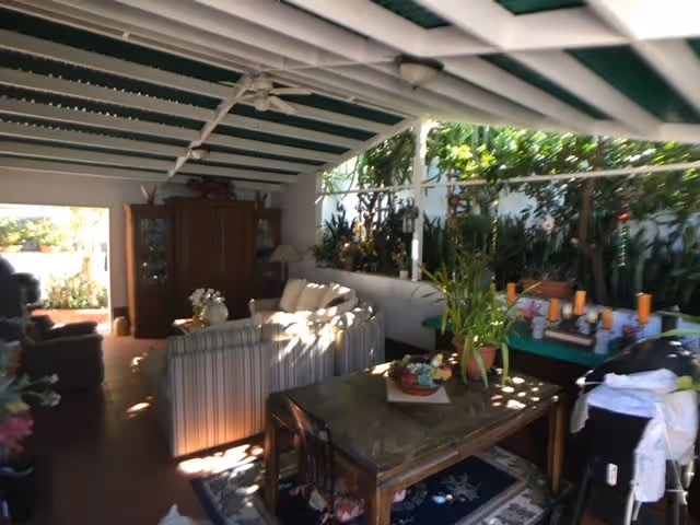 A covered outdoor patio area with a white ceiling and ceiling fan. The space includes a striped sofa, a wooden cabinet, a table with plants and decorative items, and various potted plants along the perimeter. Sunlight filters through the open sides, illuminating the seating and greenery.