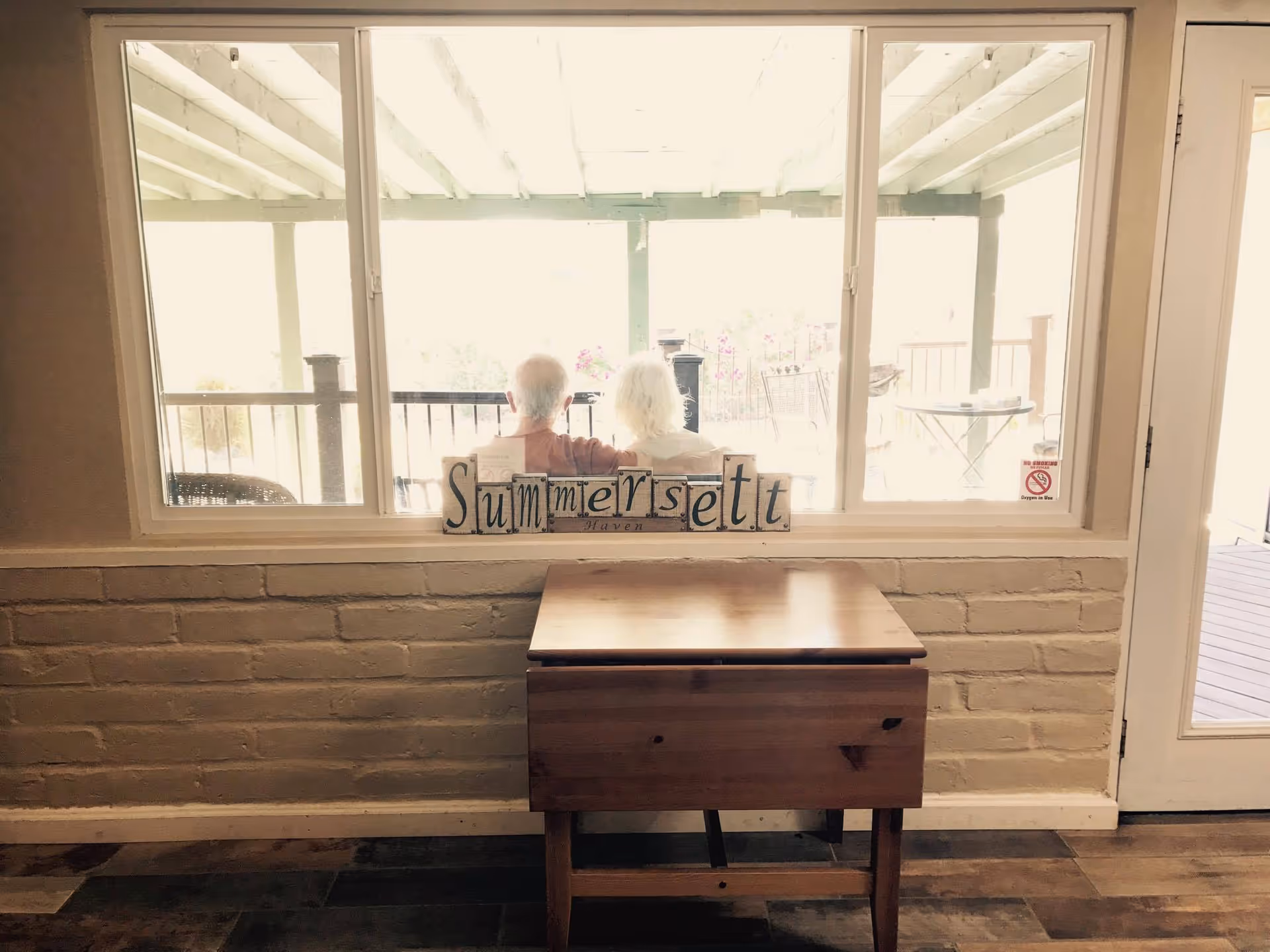 A small wooden table sits against a brick interior wall under a large window looking out to a covered porch where two people sit, with a decorative "Summersett" sign on the windowsill.