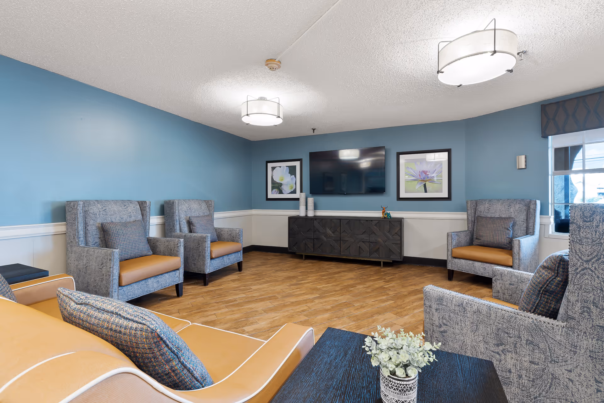 A cozy living room area with blue walls and wood flooring, featuring several upholstered armchairs and a sofa arranged around a dark wooden coffee table with a small potted plant. A flat-screen TV is mounted on the wall above a dark wooden cabinet, flanked by two framed floral pictures. Two ceiling lights illuminate the room, and a window with a patterned valance lets in natural light.