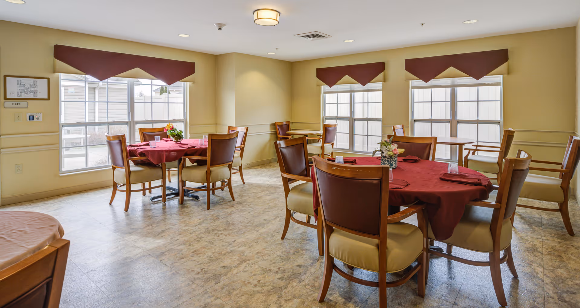 A dining room with several round tables covered with red tablecloths, each surrounded by wooden chairs with cushioned seats and backs. The room has large windows with maroon valances, beige walls, and a light-colored floor. Small flower arrangements and place settings are on the tables.