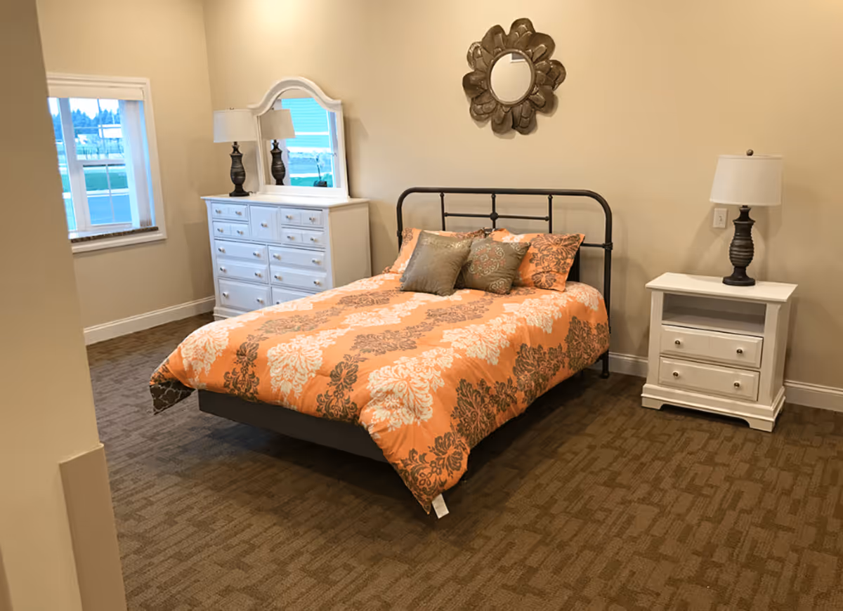 Furnished bedroom with a metal-frame bed covered in an orange patterned comforter, two white nightstands with lamps, a white dresser with mirror, and a decorative wall mirror.