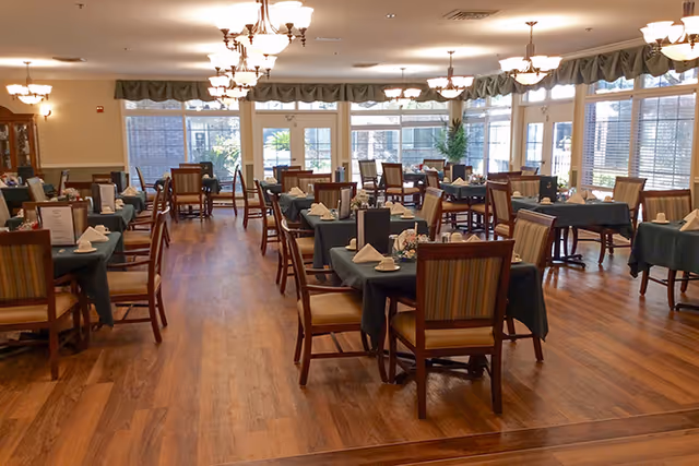 A spacious dining room with multiple tables covered in dark green tablecloths, each set with napkins and tableware. The room has wooden flooring, large windows letting in natural light, and several chandeliers hanging from the ceiling.