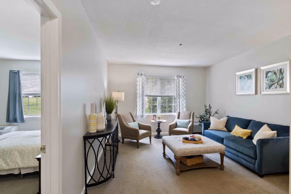 A cozy living room with a blue sofa, two beige armchairs, a large ottoman coffee table, and a small round side table near a window with patterned curtains. There are two framed artworks on the wall above the sofa, a floor lamp, and decorative vases on a console table along the left wall. A bedroom with a bed and blue curtains is visible through an open doorway on the left.