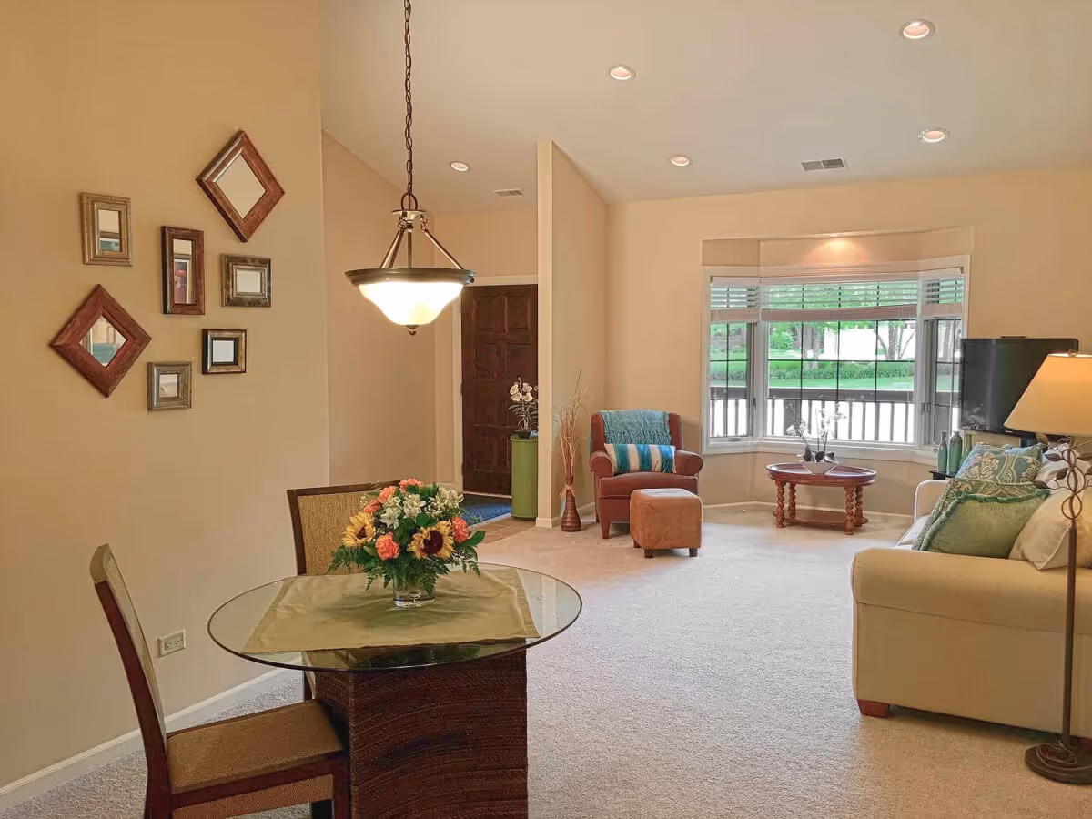 A cozy living room and dining area in an independent senior living facility. The room features a round glass dining table with two chairs and a floral centerpiece. The living area includes a beige sofa with green and white pillows, a brown armchair with a matching ottoman, a wooden coffee table, a floor lamp, and a TV near a large window with white blinds. The walls are decorated with a set of small framed mirrors, and the room is softly lit with recessed ceiling lights and a hanging light fixture above the dining table.