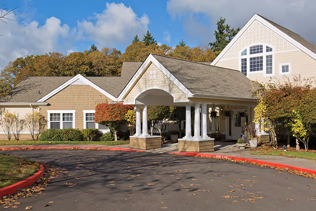 Exterior view of a senior living facility with a covered entrance supported by white columns. The building has beige siding and multiple windows, surrounded by trees with autumn foliage under a partly cloudy sky.