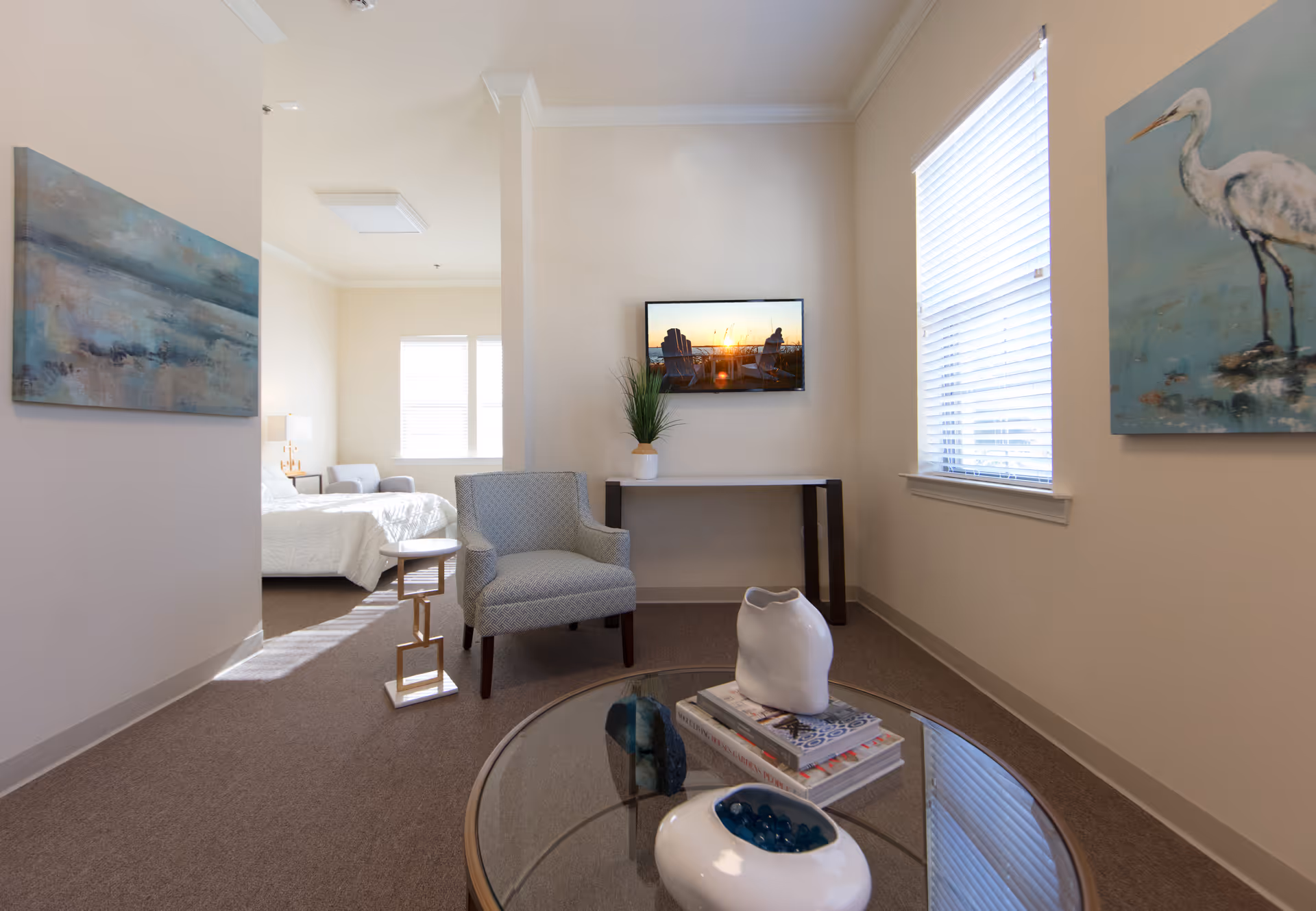 A bright and cozy living area with a light blue armchair, a glass coffee table with decorative items and books, and a wall-mounted TV displaying a sunset scene. The room has beige walls with two paintings, one of a bird and another abstract landscape. A bedroom with a white bed and armchair is visible through an open doorway.