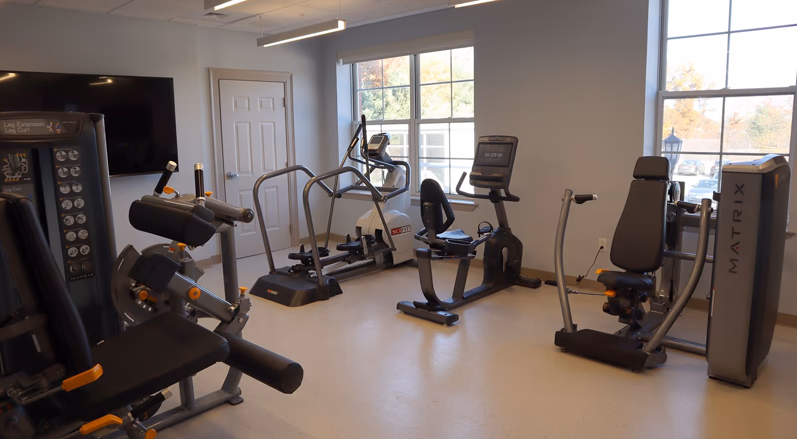 A well-lit exercise room with various fitness equipment including a leg extension machine, a recumbent bike, an elliptical trainer, and a seated chest press machine. The room has large windows letting in natural light and a wall-mounted television.