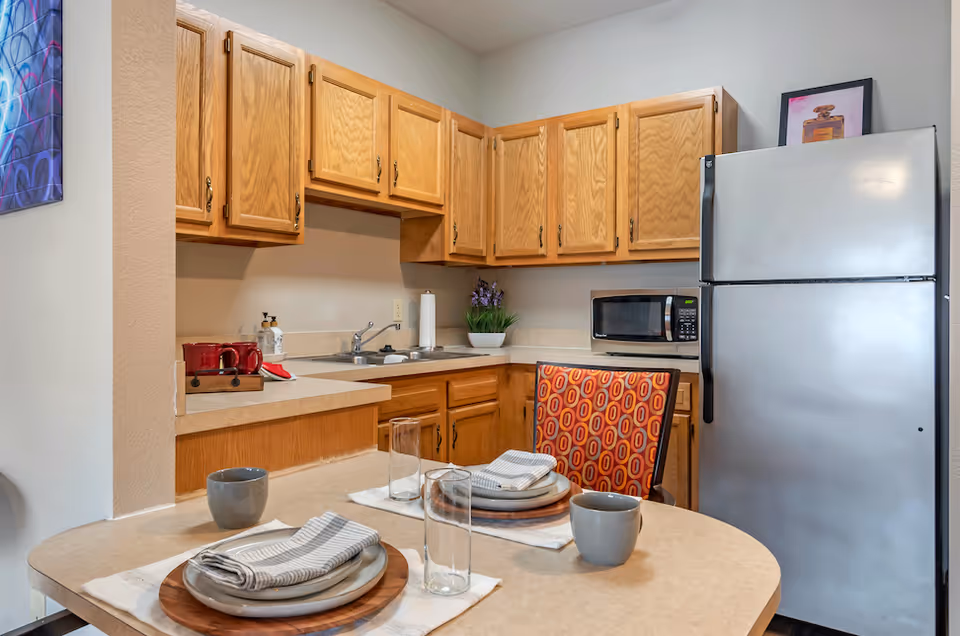 A small kitchen area with wooden cabinets, a stainless steel refrigerator, a microwave, and a sink. In the foreground, there is a dining table set for two with plates, napkins, glasses, and mugs. One chair with a red and orange patterned cushion is visible. A small plant and a framed picture are on the countertop.