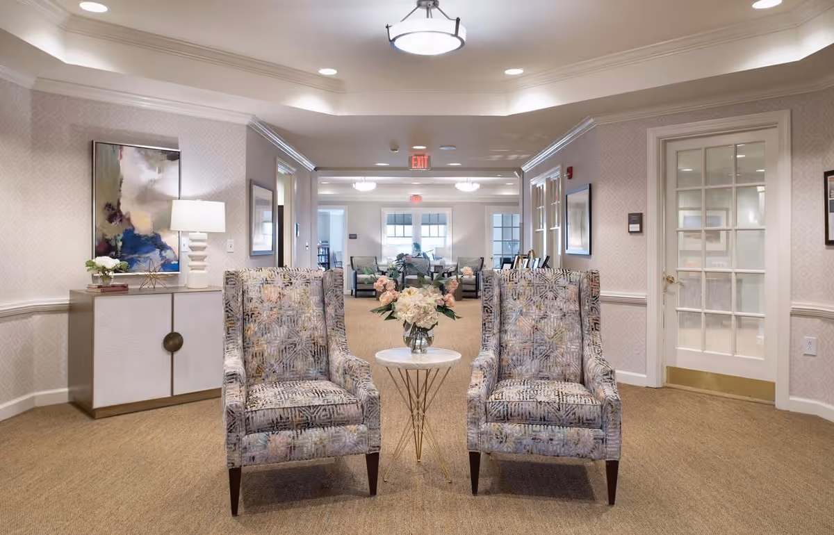 A bright and elegant sitting area in a senior living facility featuring two patterned armchairs with a small round table holding a vase of flowers between them. The room has beige carpet, light-colored wallpaper, a sideboard with a lamp and artwork, and a glass-paneled door on the right. In the background, there is another seating area with chairs and tables near large windows.