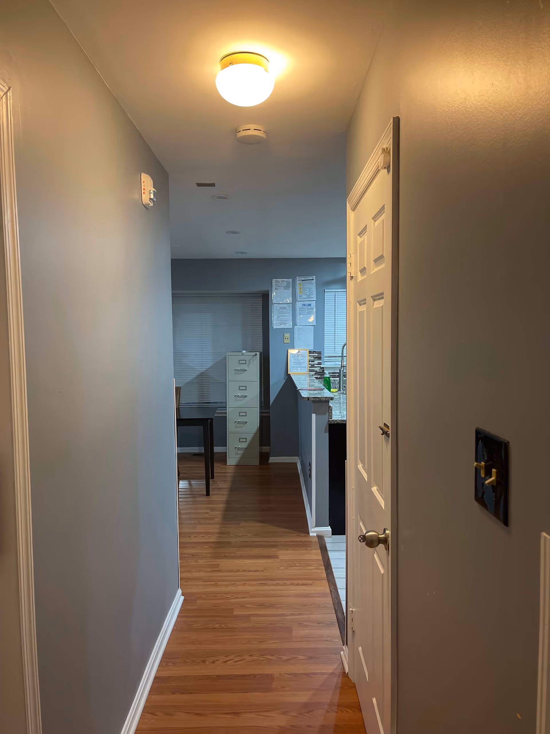 A hallway with wooden flooring leading to a room with a filing cabinet and a table. The walls are painted gray, and there is a ceiling light fixture illuminating the hallway. On the right side, there is a closed white door and a light switch on the wall.