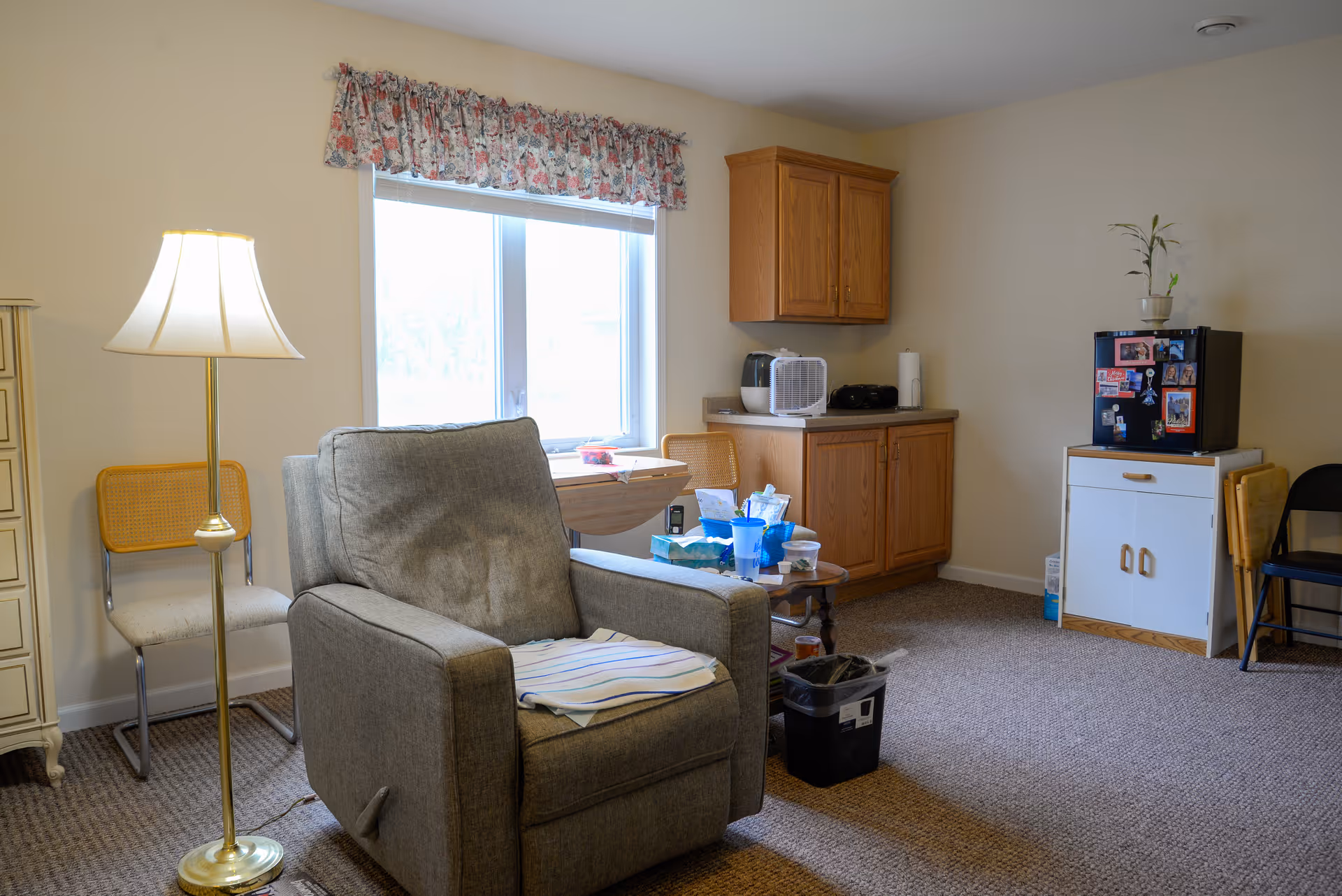 A cozy living area with a gray recliner chair, a floor lamp, and a window with floral valance. There are two chairs against the wall, a small wooden table with various items, a cabinet with a microwave and paper towel, and a small refrigerator with photos and magnets on it. The room has beige walls and carpeted floor.