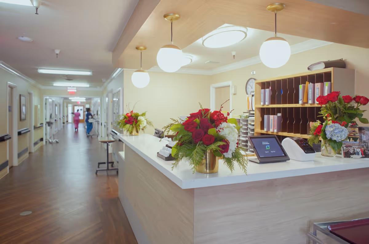 Reception desk area in a healthcare facility hallway with flower arrangements on the counter, shelves with files behind the desk, and two healthcare workers in scrubs walking down the corridor.