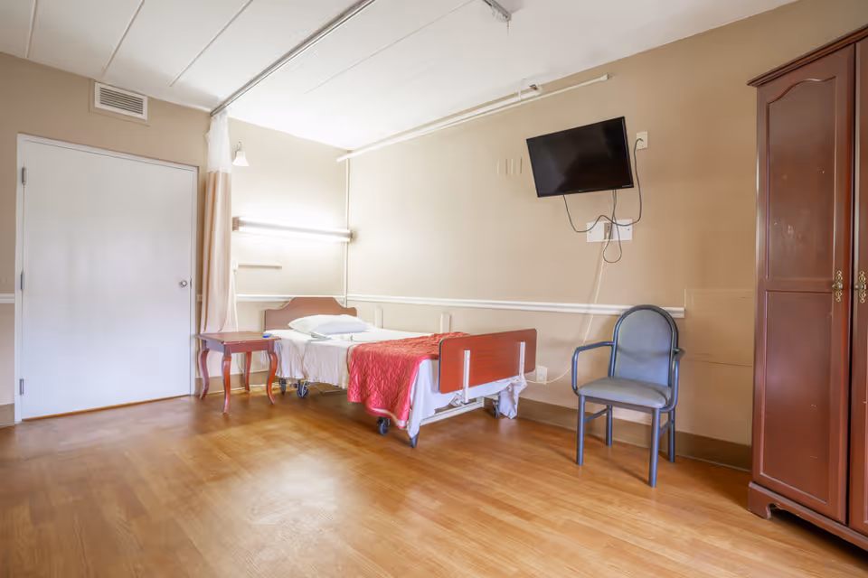 A simple nursing-home bedroom with a single bed, bedside table, wall-mounted TV, chair, and wardrobe.