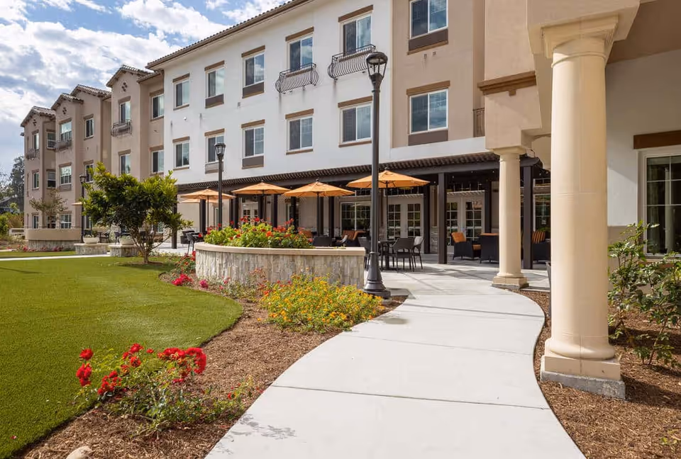 Landscaped courtyard and outdoor patio with umbrellas and seating in front of a multi-story senior living building.
