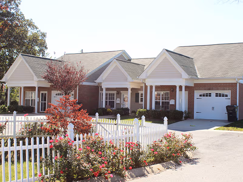 Exterior view of a single-story senior living facility building with brick and white siding, a white picket fence, flowering bushes, and a driveway leading to a garage.