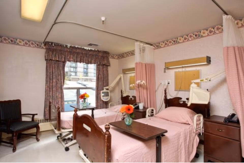 A nursing facility room with two hospital beds covered in pink bedding, each with a small table holding a vase with orange flowers. The room has floral wallpaper border, a window with floral curtains, a wooden chair, and medical equipment mounted on the walls. Privacy curtains hang between the beds.