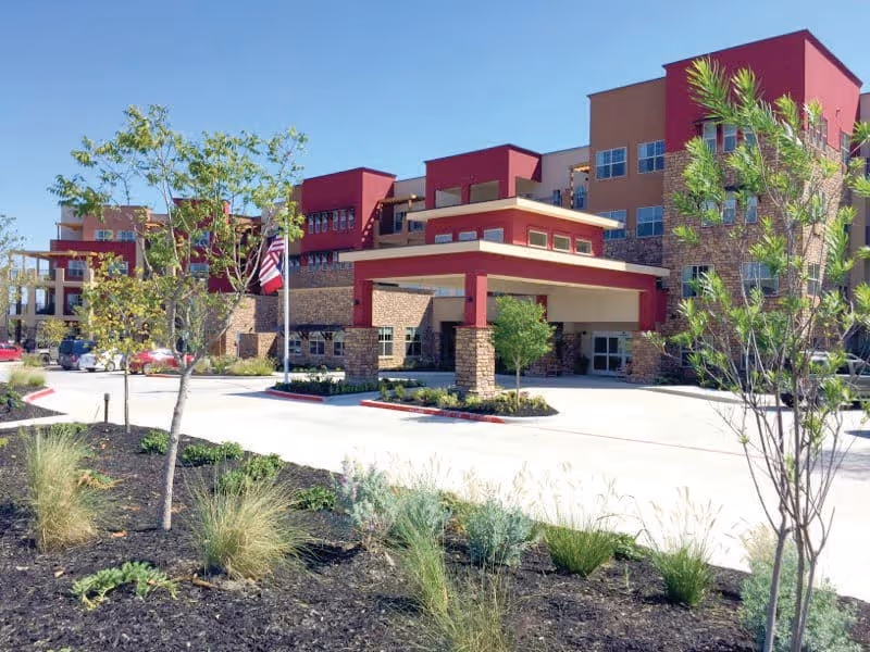 Exterior view of a modern multi-story senior living facility with a covered entrance, landscaped garden with small trees and shrubs, and a clear blue sky.