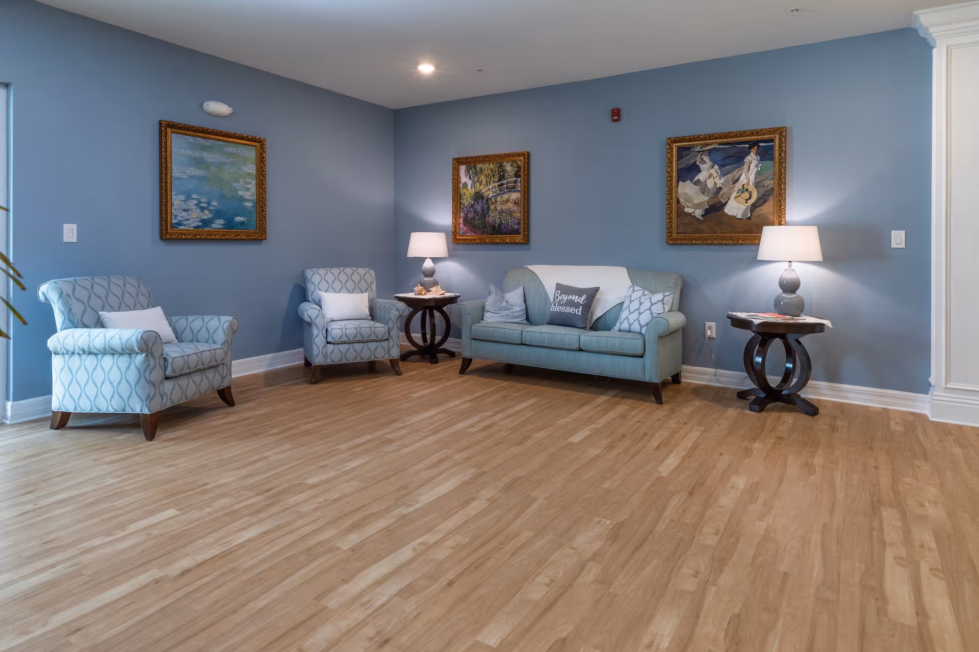 A light-blue seating area with two patterned armchairs and a sofa arranged against a blue wall, side tables with lamps, and framed paintings.