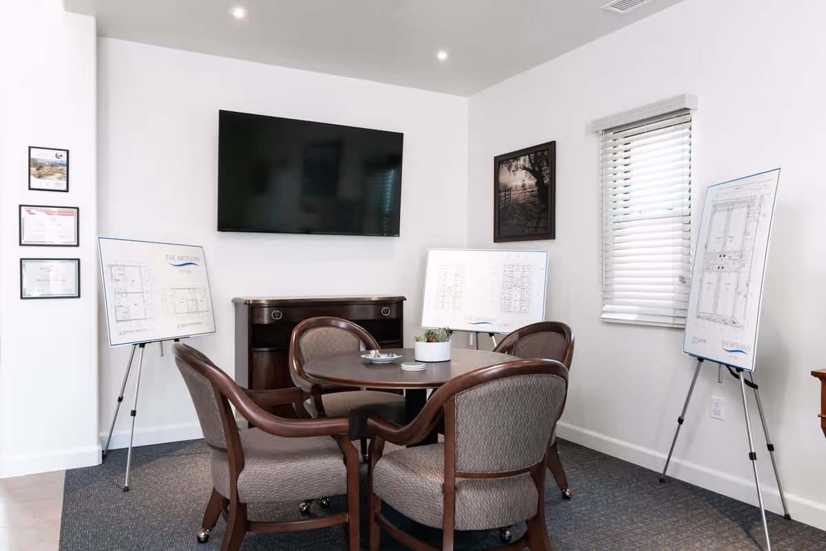 A small meeting room with a round wooden table surrounded by four cushioned chairs. On the table is a small plant and some coasters. The room has a wall-mounted flat screen TV, framed certificates on one wall, a framed black and white photo on another wall, and three easels displaying architectural floor plans labeled 'The Artesian'. There is a window with white blinds partially open.