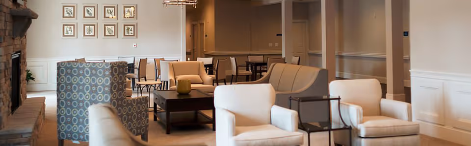 A cozy living room area in a senior living facility featuring multiple beige armchairs and sofas arranged around a dark wooden coffee table. The room has a stone fireplace on the left, framed artwork on the far wall, and several dining tables and chairs in the background. The walls are painted light beige with white wainscoting.