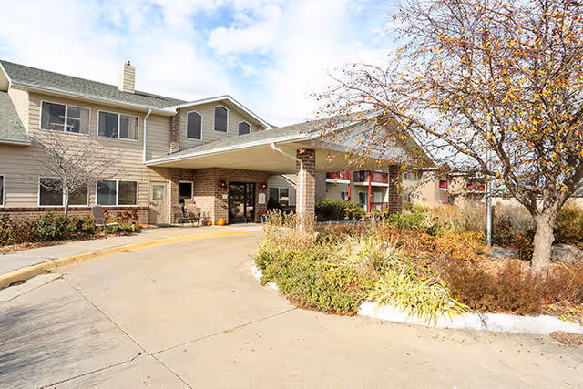 Covered front entrance of a two-story senior living building with a circular driveway and landscaped plantings.