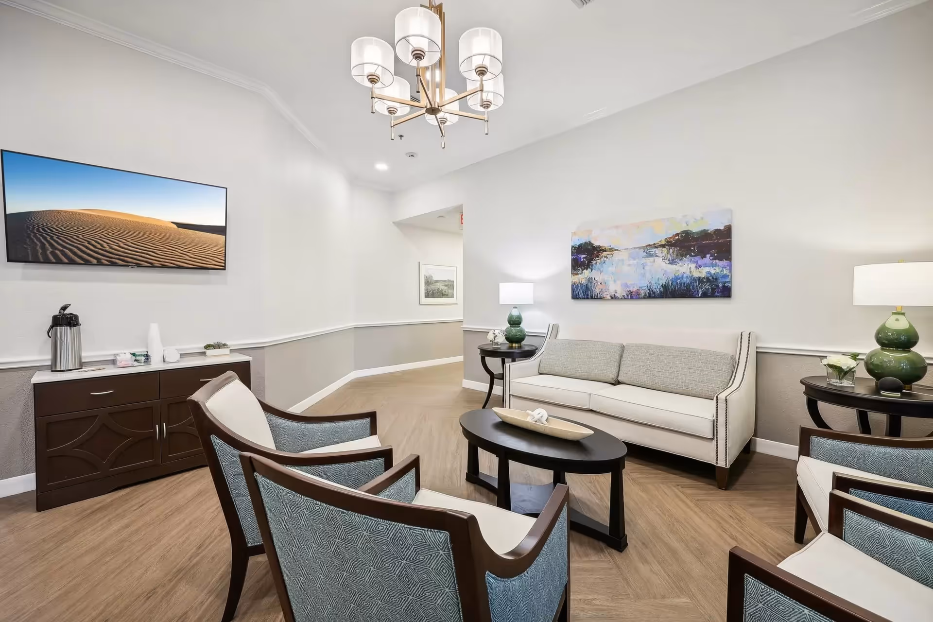 A well-lit living room with a beige sofa, four armchairs with patterned upholstery, a black oval coffee table, two side tables with green lamps, a wall-mounted TV displaying a desert landscape, and a colorful landscape painting on the wall.