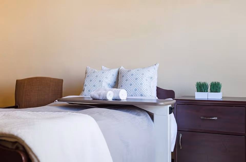 A furnished bedroom with a bed topped by decorative pillows, an overbed table holding rolled towels, and a bedside dresser with small potted greenery.