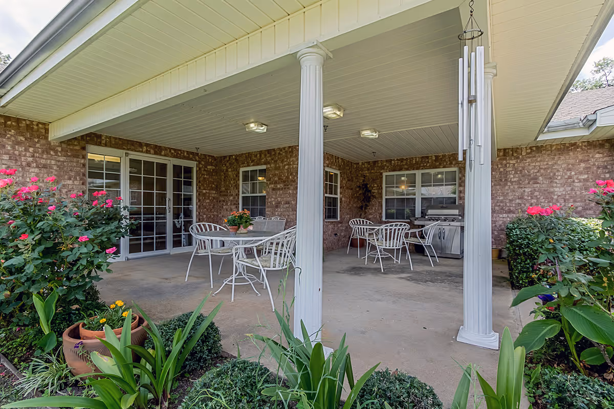 Covered outdoor patio area with white columns, several white metal tables and chairs, potted plants on the tables, a stainless steel grill, and surrounding garden with green plants and pink flowers.