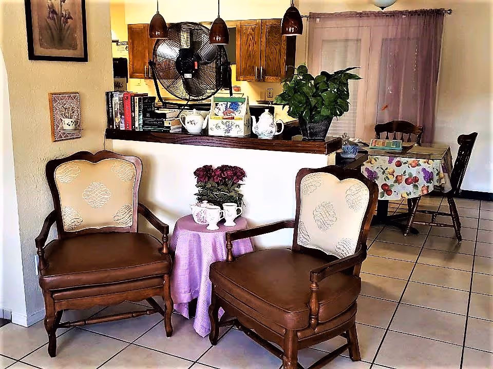 A cozy interior space featuring two wooden armchairs with beige patterned cushions placed around a small round table covered with a purple cloth. On the table are two white mugs and a small potted plant with purple flowers. Behind the chairs is a counter with wooden cabinets above, a large black fan, books, and decorative items including a teapot and a plant. In the background, there is a dining table covered with a fruit-patterned tablecloth and wooden chairs, with sheer purple curtains on the window.