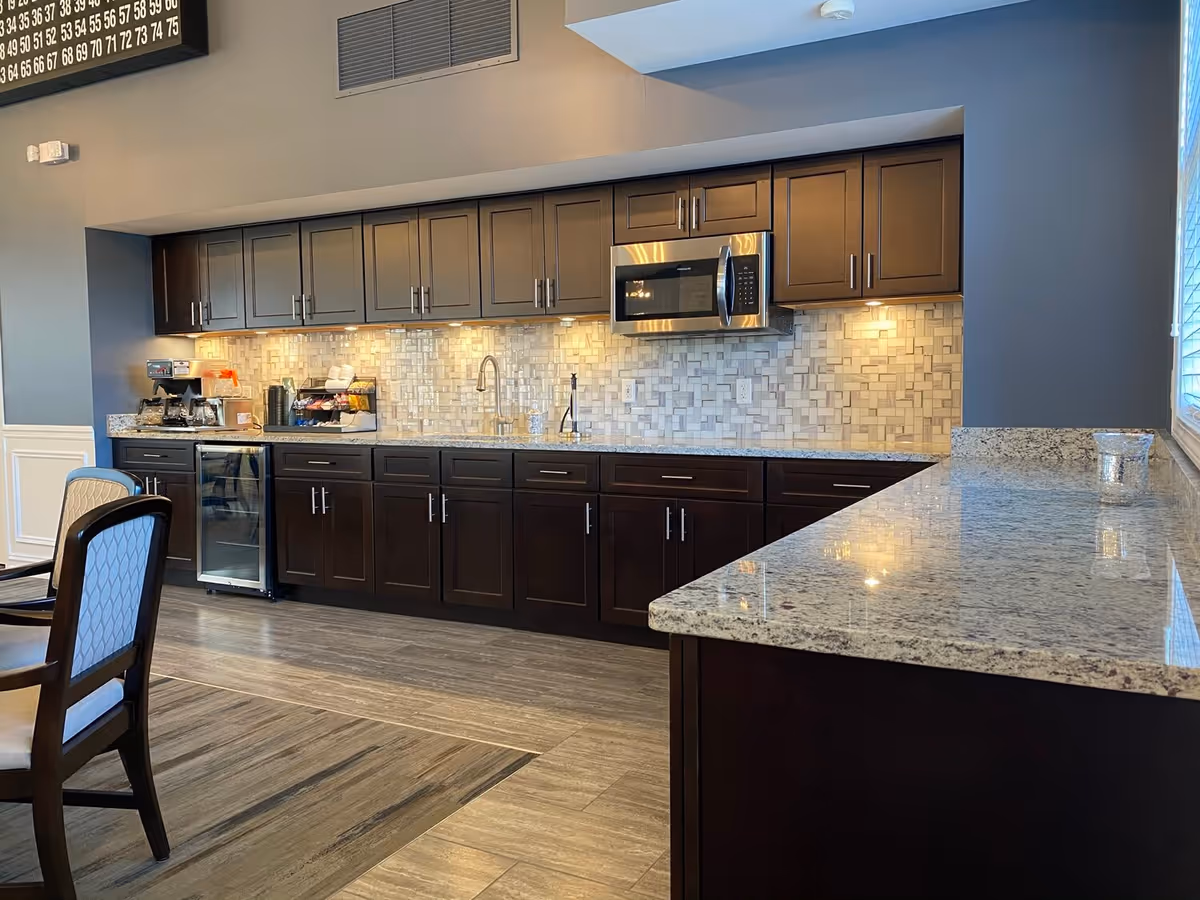 A modern kitchen area in a senior living facility featuring dark wood cabinets, a granite countertop, a built-in microwave, a small refrigerator, a coffee maker, and a tiled backsplash. There are chairs with patterned upholstery partially visible on the left side and a large window on the right side letting in natural light.