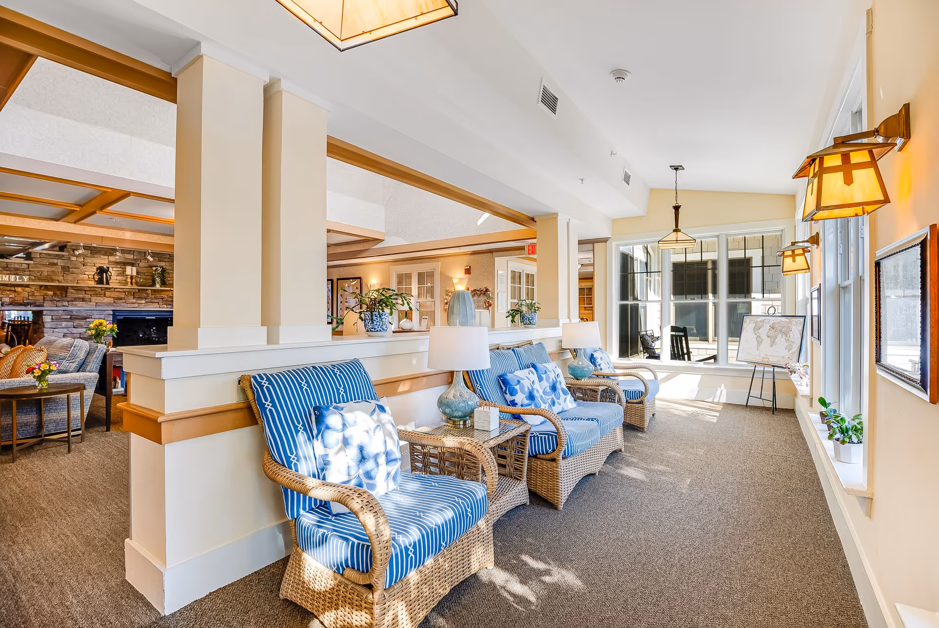 A bright and cozy sitting area in a senior living facility with wicker chairs featuring blue and white cushions, small side tables with lamps, and large windows letting in natural light. The space is decorated with plants and wall-mounted lights, with a view into an adjacent room with a stone fireplace and more seating.